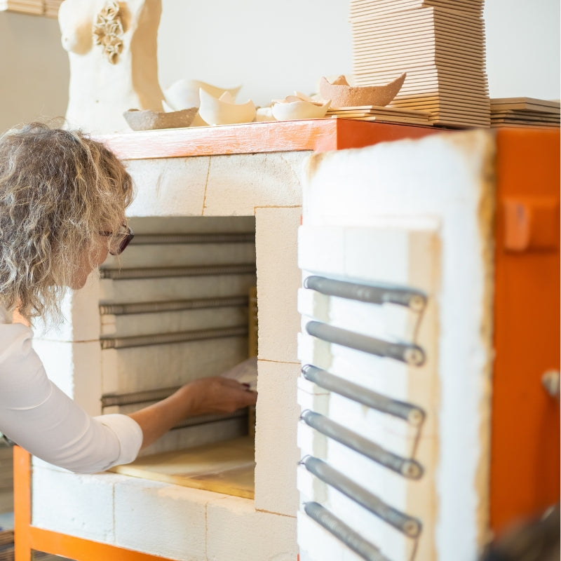 A woman placing pottery inside a kiln, surrounded by ceramic pieces and stacked tiles in a workshop setting